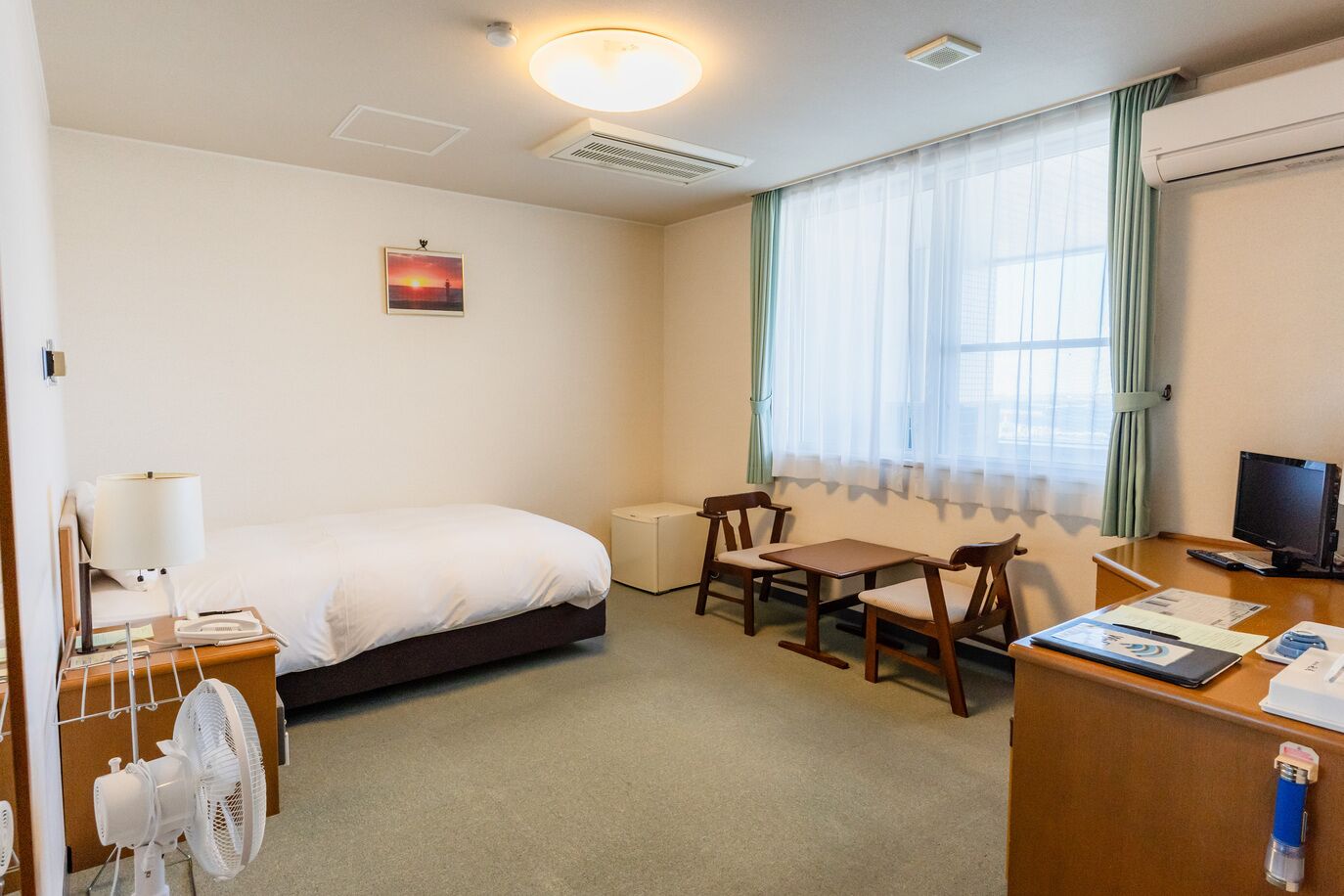 A hotel room with a bed, a desk with a computer, a chair, a fan, and a window with curtains.