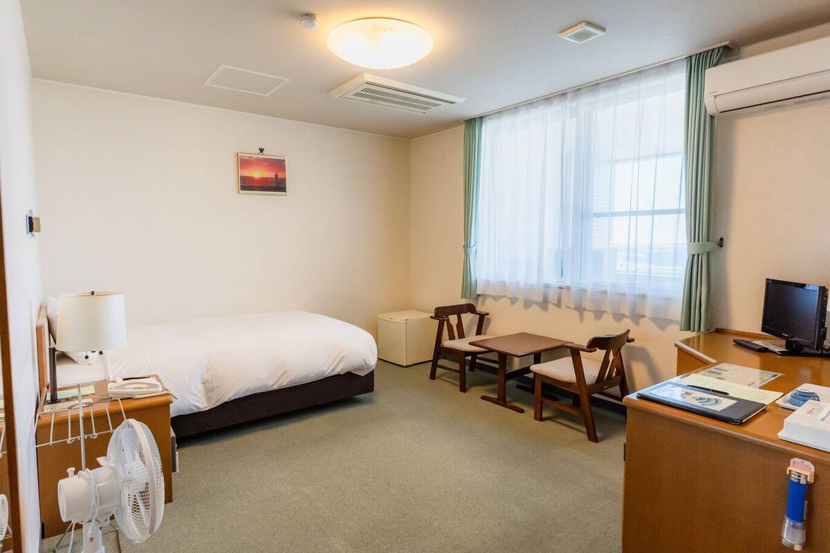 A hotel room with a bed, a desk with a computer, a chair, a fan, and a window with curtains.