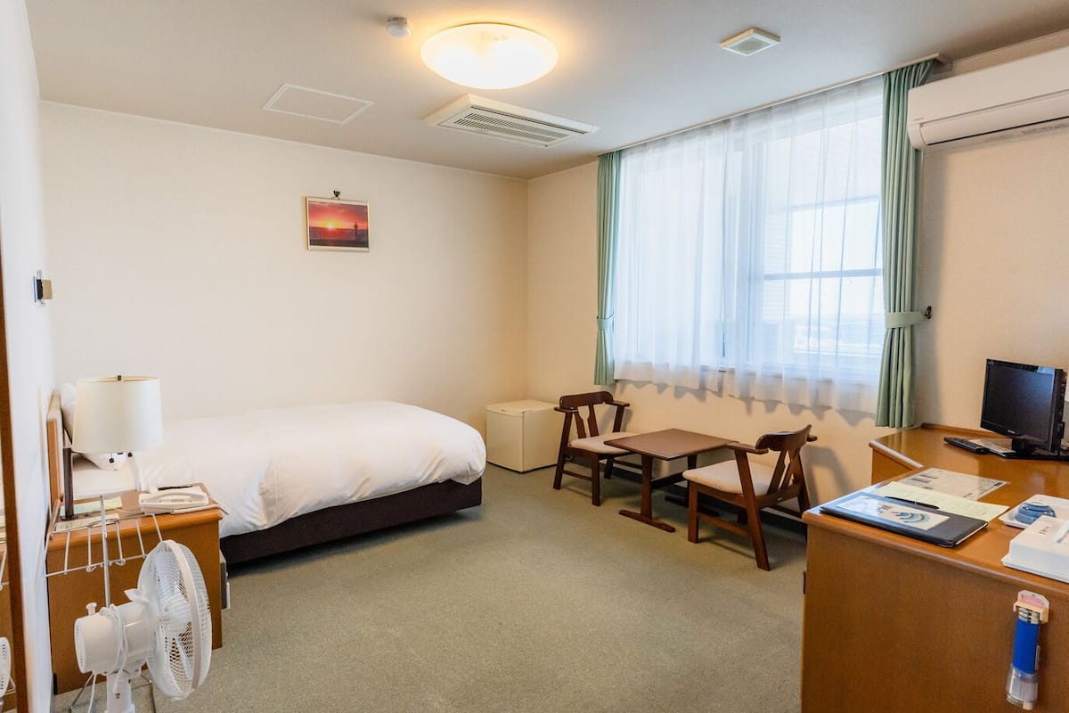 Une chambre d’hôtel avec un lit, un bureau équipé d’un ordinateur, une chaise, un ventilateur et une fenêtre avec des rideaux.