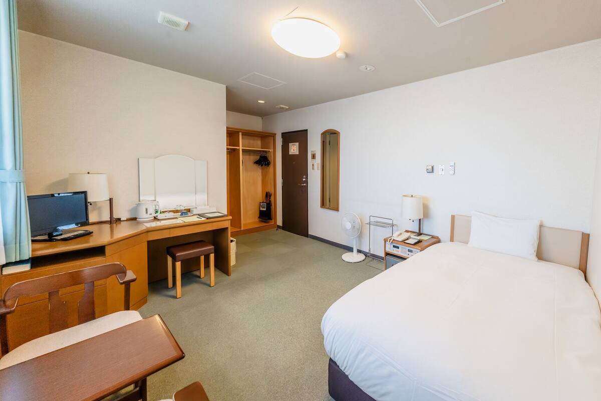 A hotel room with a bed, desk, chair, and a small table with a lamp.