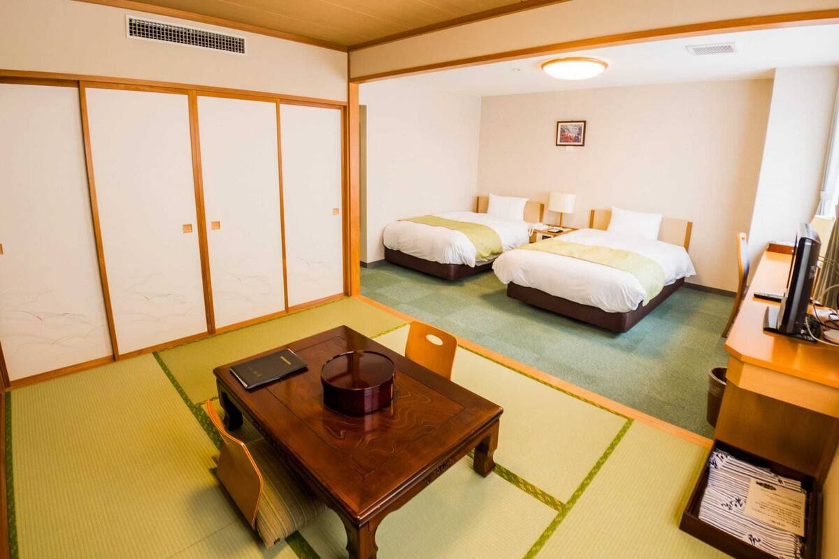 A hotel room with two beds, a dining table, a TV, and a traditional Japanese tatami mat floor.