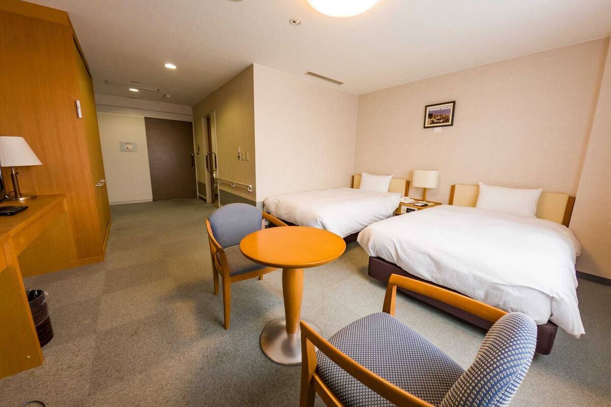 A hotel room with two beds, a desk with a chair, a small round table, and a door leading to another room.