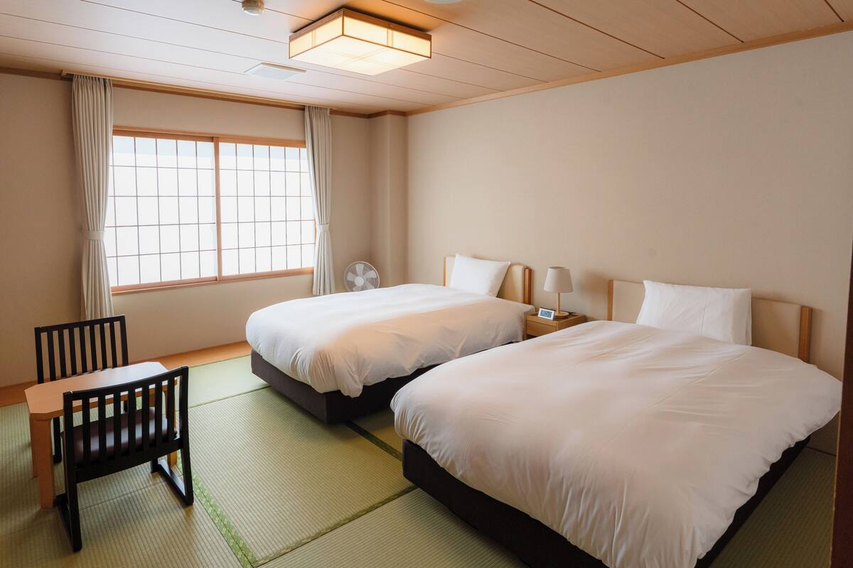 A hotel room with two beds, a small table, a chair, a fan, and a window with curtains.