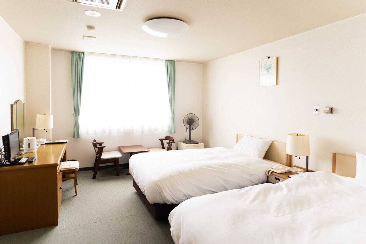 A hotel room with two beds, a desk with a chair, a bedside table with a lamp, a fan, and a window with curtains.
