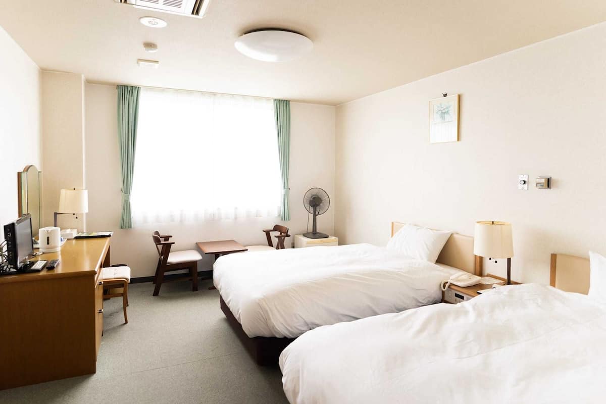 Une chambre d’hôtel avec deux lits, un bureau avec une chaise, une table de chevet avec une lampe, un ventilateur et une fenêtre avec des rideaux.