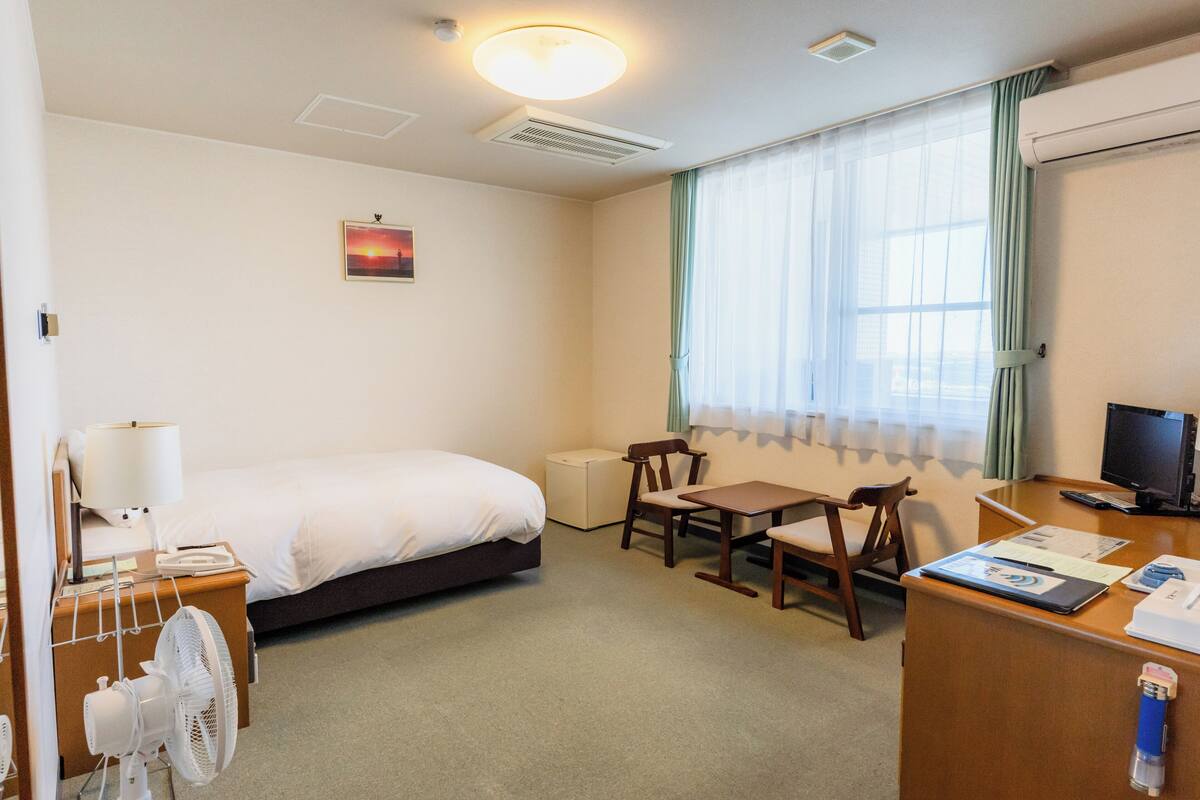 A hotel room with a bed, desk, chair, fan, and a window with curtains.