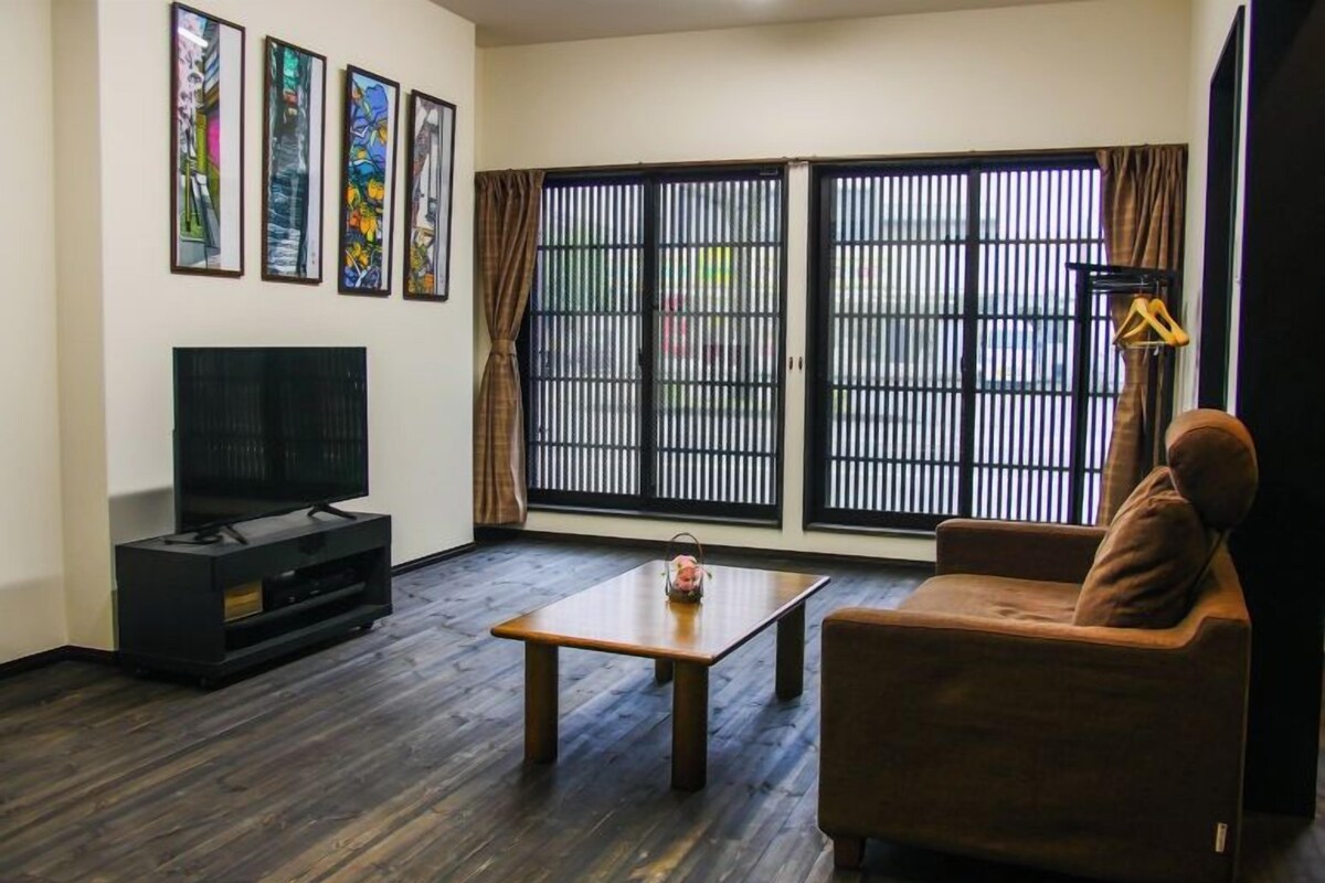 A living room with a flat-screen TV, a coffee table, a sofa, and a large window with blinds.