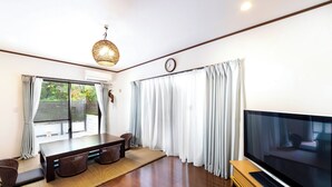 A modern living room with a large flat-screen TV, a dining table with chairs, and a large window with curtains. - Sea Stay Okinawa (Onna)