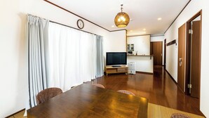 A modern living room with a wooden dining table, wicker chairs, a flat-screen TV, and a kitchen area in the background. - Sea Stay Okinawa (Onna)