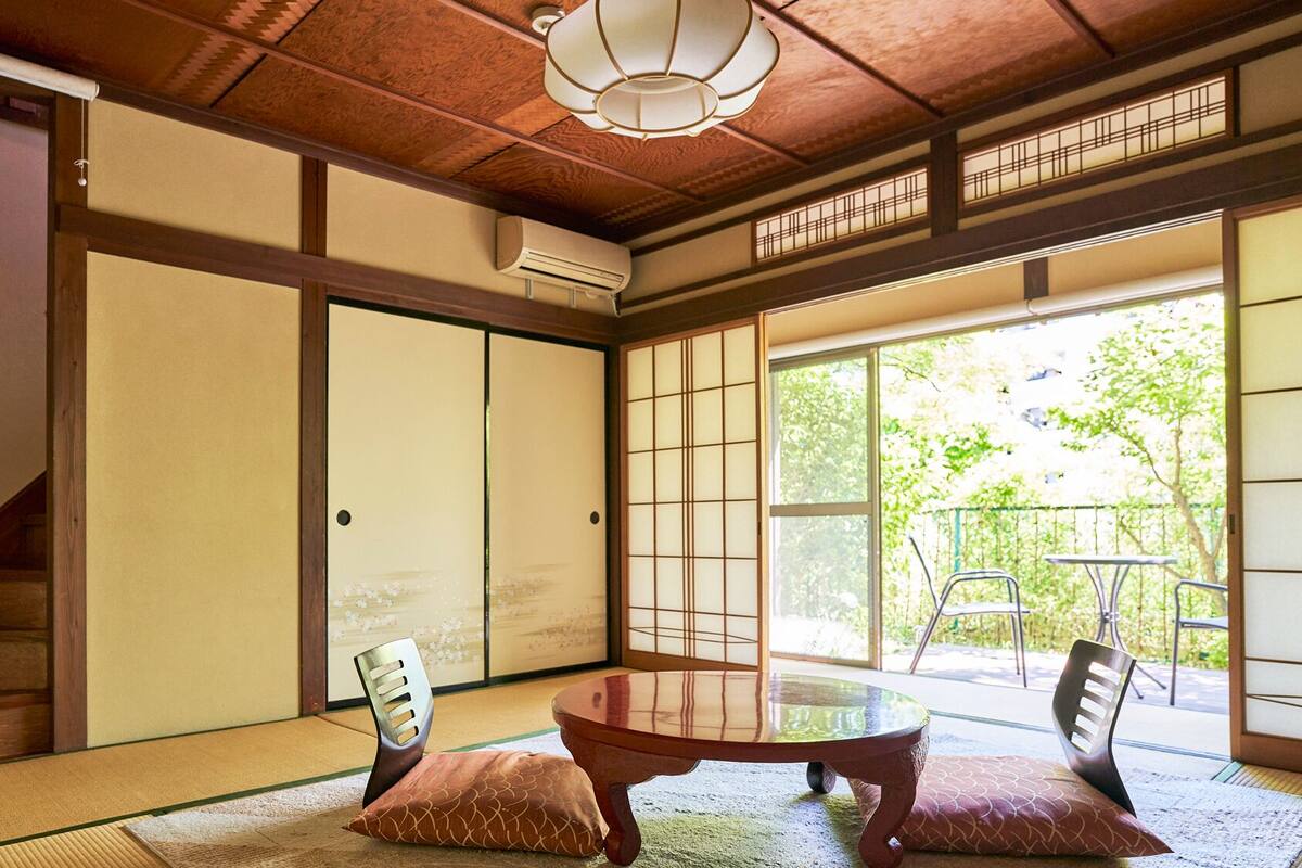 Une pièce de style japonais traditionnel, avec un sol recouvert de tatamis, des portes coulissantes, une table avec des coussins et une vue sur la verdure à l’extérieur.