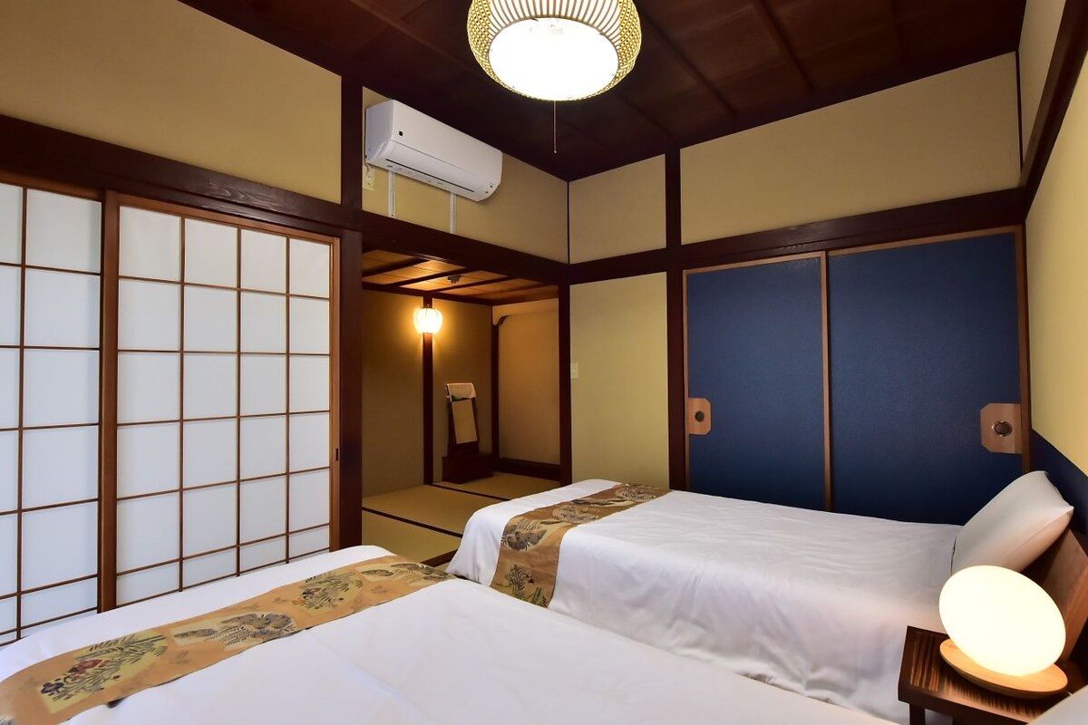 Una habitación japonesa tradicional con suelo de tatami, dos camas con ropa de cama blanca, un ventilador de techo y un aire acondicionado instalado en la pared.
