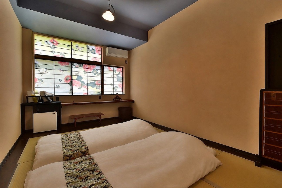 Habitación de hotel con dos camas, un escritorio, una silla, una mesita y un radiador. Se ve una ventana con motivos florales.