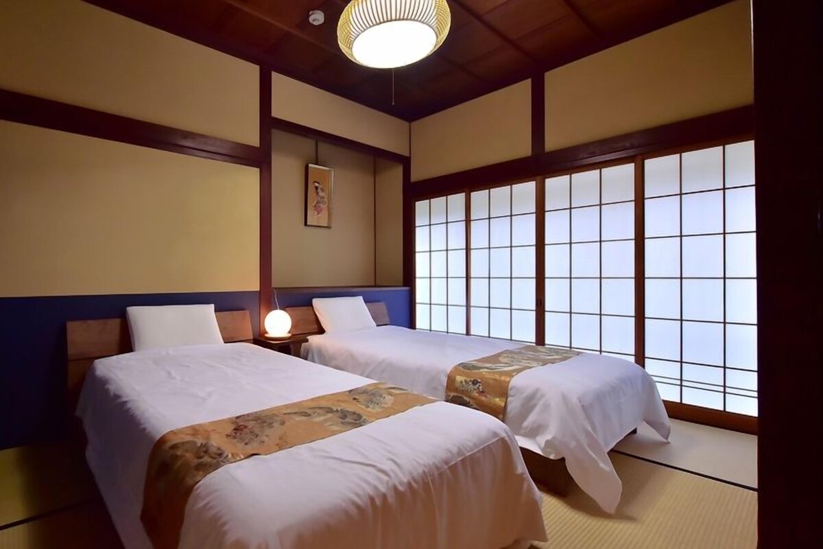 Une pièce de style japonais traditionnel, avec un sol recouvert de tatamis, deux lits avec du linge de lit blanc, un paravent shoji suspendu et un luminaire au plafond.