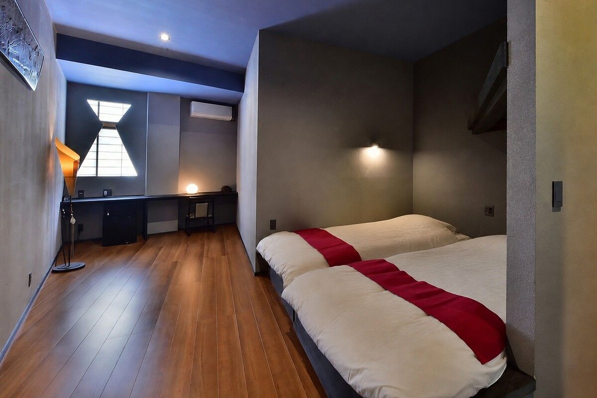 Habitación de hotel con dos camas, un escritorio, una silla y una ventana con lámpara de diseño geométrico.