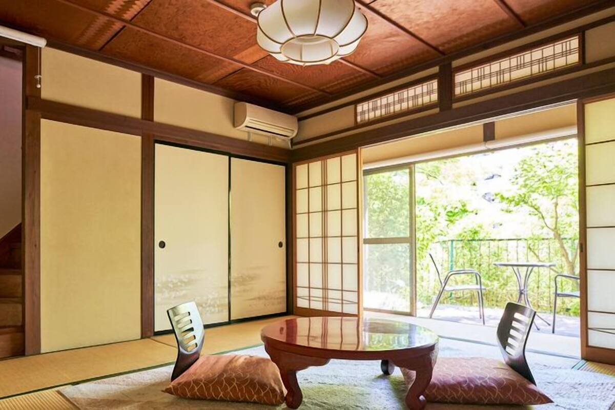 Une pièce de style japonais traditionnel, avec un sol recouvert de tatamis, des portes coulissantes, une petite table basse avec des coussins et une vue sur la verdure à l’extérieur.