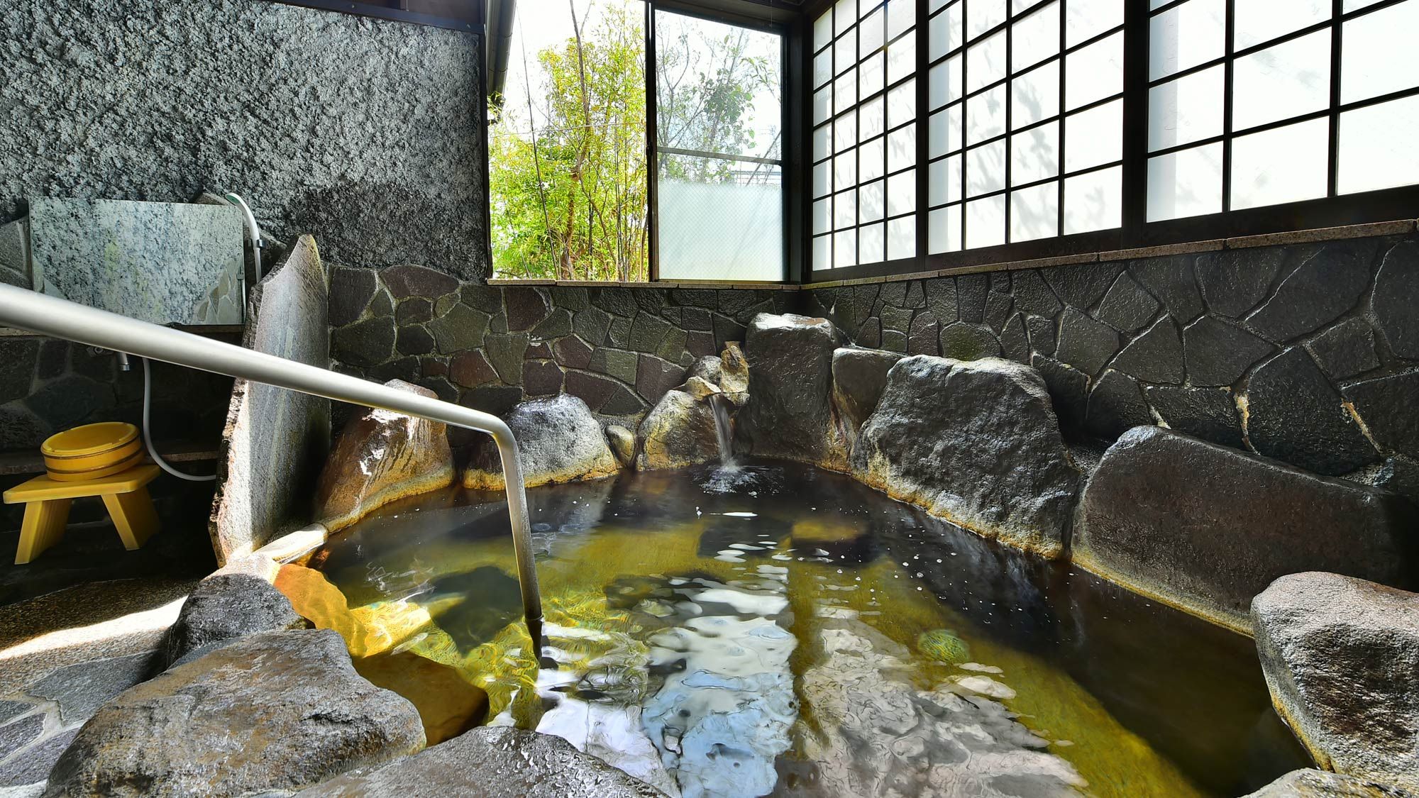 A hot spring bath with a stone wall, a wooden stool, and a metal railing.