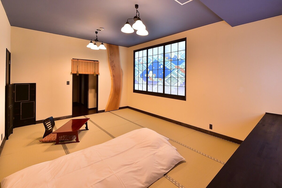 Una habitación con suelo de tatami, un futón, una mesita baja, un televisor y un ventanal con vistas.