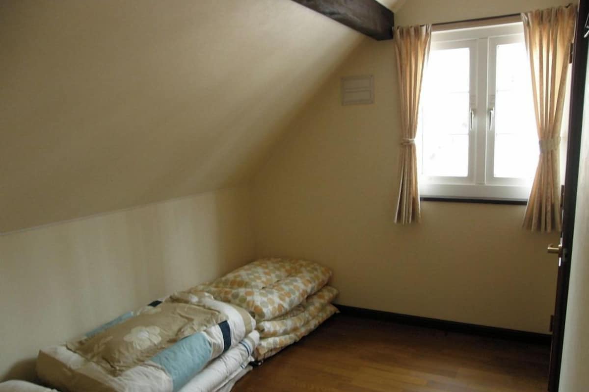 Um quarto pequeno e vazio, com teto inclinado, janela com cortinas e piso de madeira.