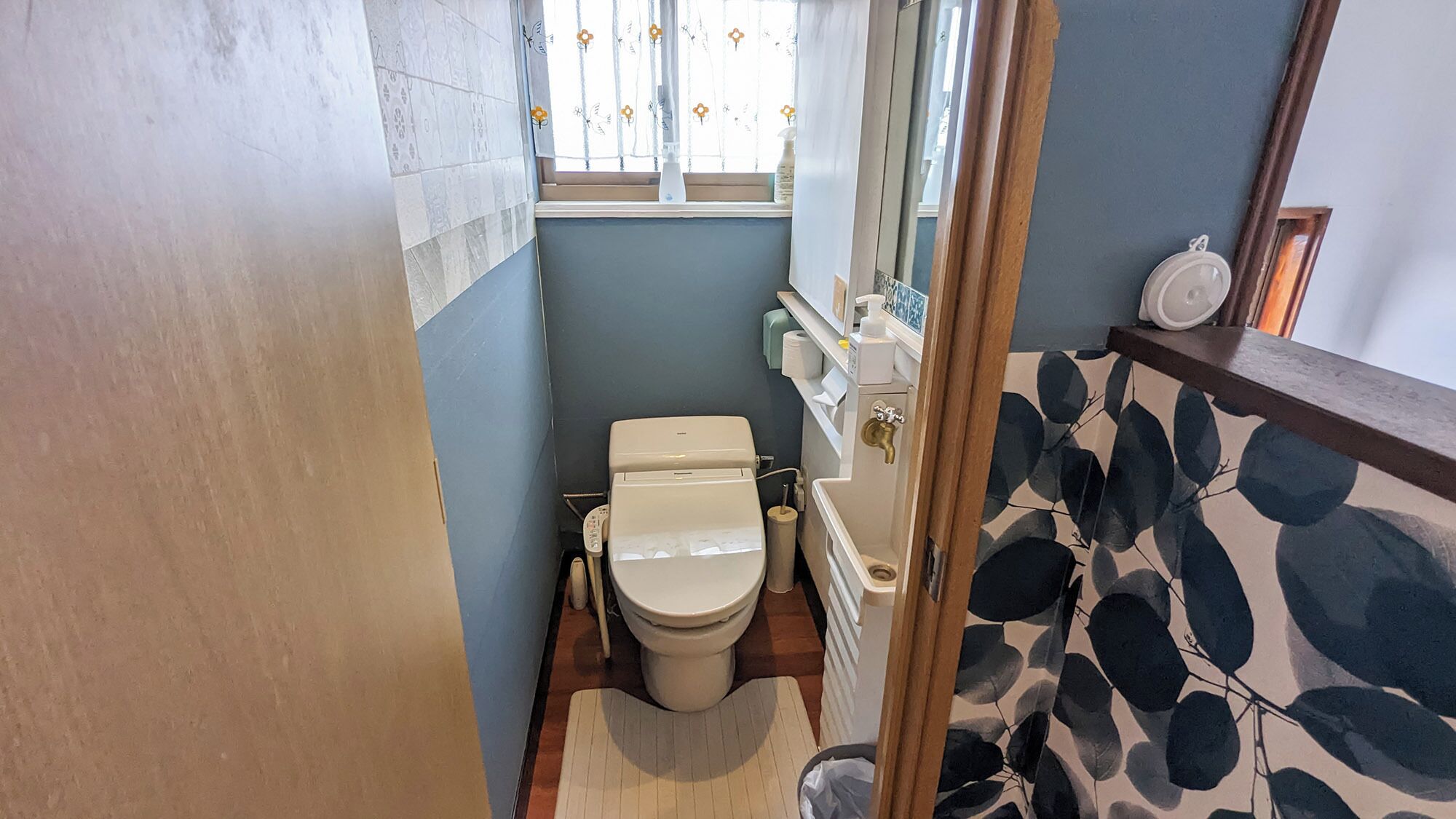 A compact bathroom with a toilet, a sink, and a window with floral curtains.
