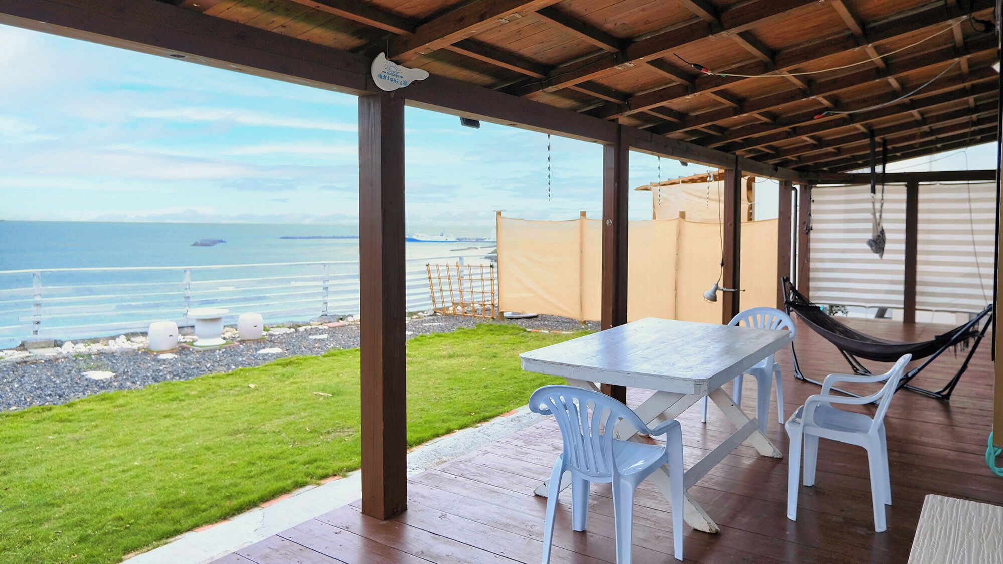 Een overdekt terras met een tafel en stoelen, met uitzicht op een strand en de oceaan, met een schip in de verte.