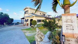 A two-story house with a large front lawn, a palm tree, and a sign indicating it's a cottage. - Sea Turtle View Cottage (Wadomari)