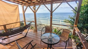 A wooden deck with a hammock, wicker chairs, and a glass table overlooking the ocean. - Sea Turtle View Cottage (Wadomari)