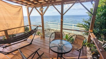 Una terraza de madera con una hamaca, sillas de mimbre y una mesa de vidrio con vistas al océano.