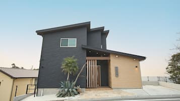 A modern two-story building with a dark facade, a wooden entrance, and a small garden area with plants.