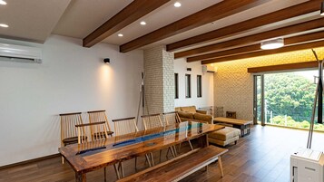 Un espace de repas moderne avec une longue table en bois, plusieurs chaises, un canapé et une vue sur la verdure grâce à une grande fenêtre.