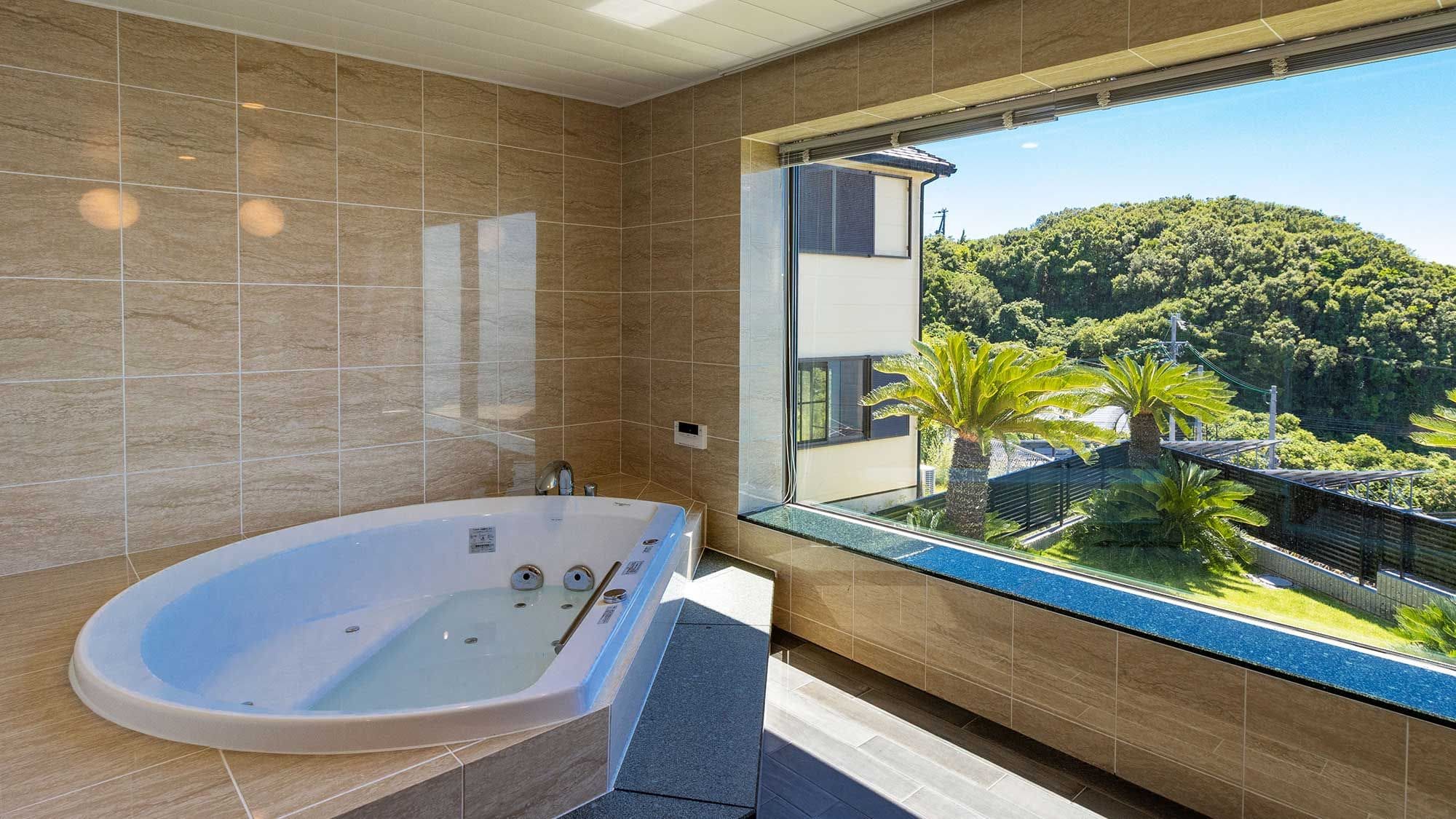 A modern bathroom with a large bathtub, tiled walls, and a balcony with a view of a lush green hill.