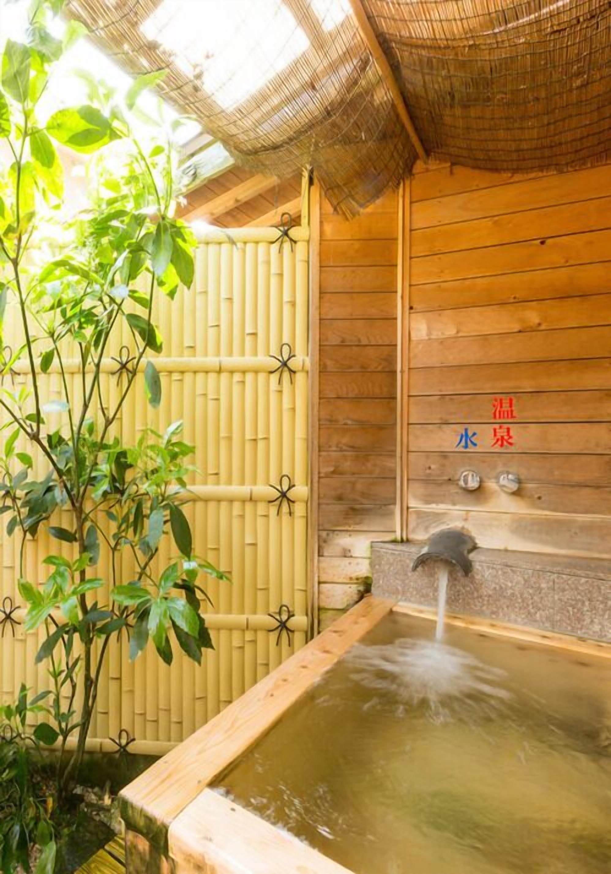 A traditional wooden bathhouse with a bamboo fence, a water spout, and a sign indicating temperature control.