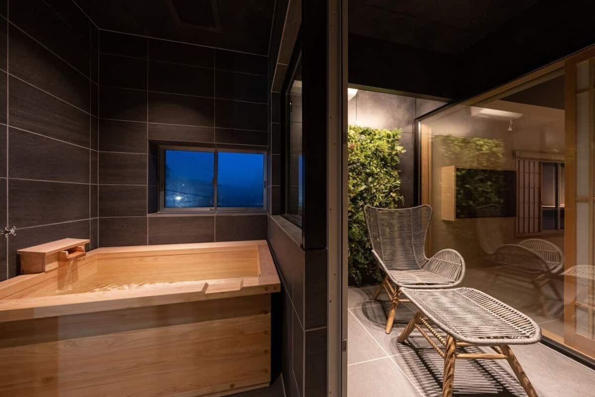 A modern bathroom with a wooden bathtub, dark tiled walls, a window with a view, and wicker chairs outside.