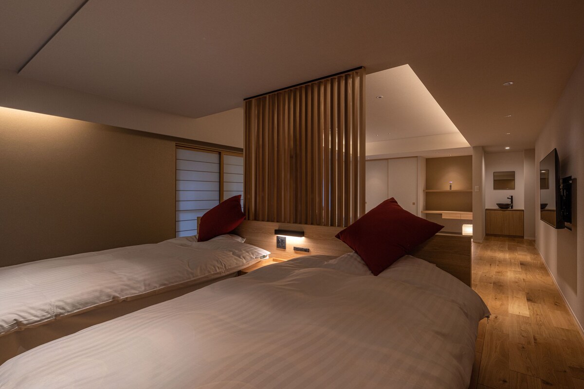 A modern hotel room with two beds, a wooden floor, a large window with blinds, a bedside table with a lamp, and a flat-screen TV mounted on the wall.