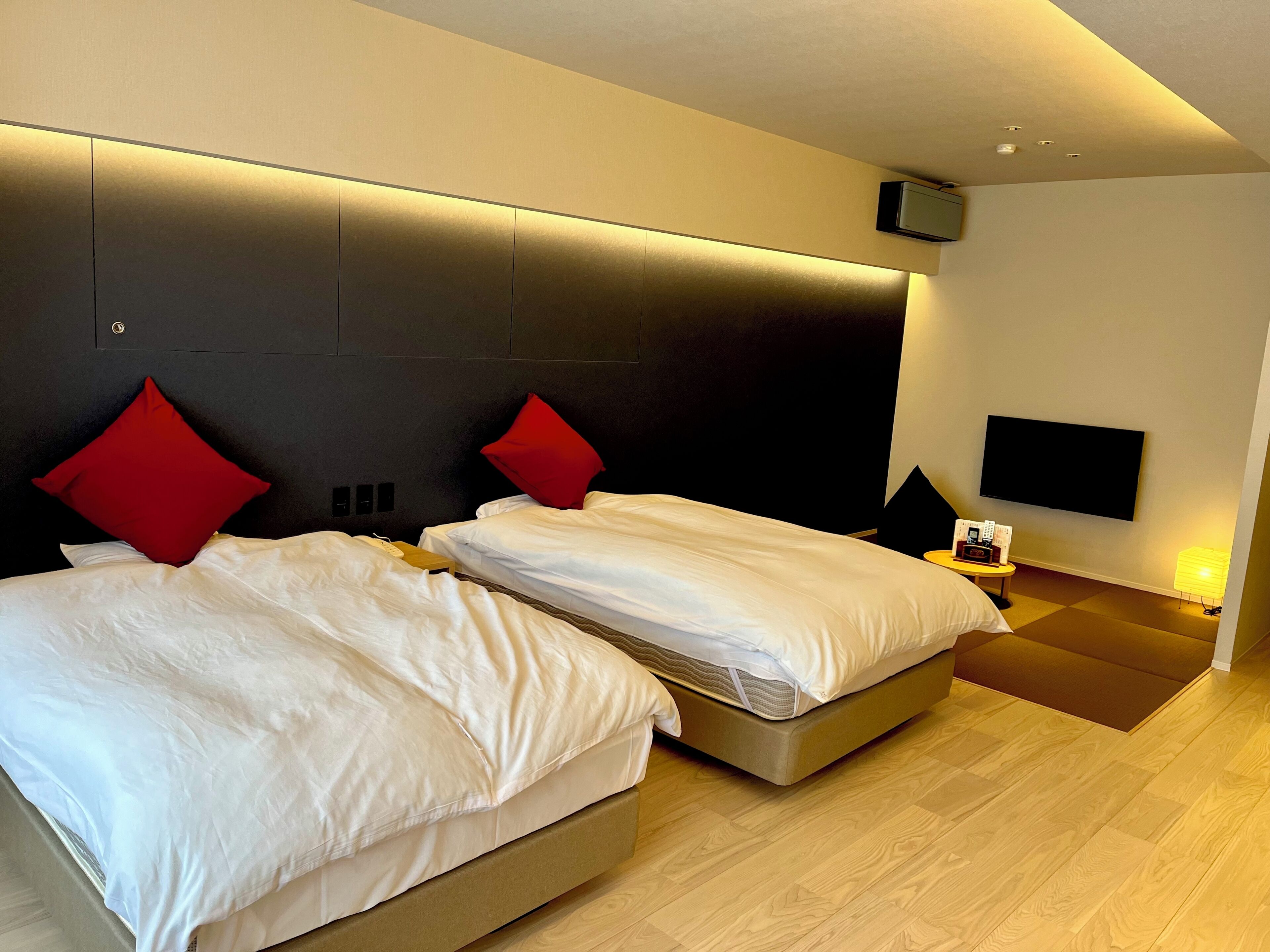 A modern hotel room with two beds, a flat-screen TV, a bedside table with a lamp, and a wooden floor.