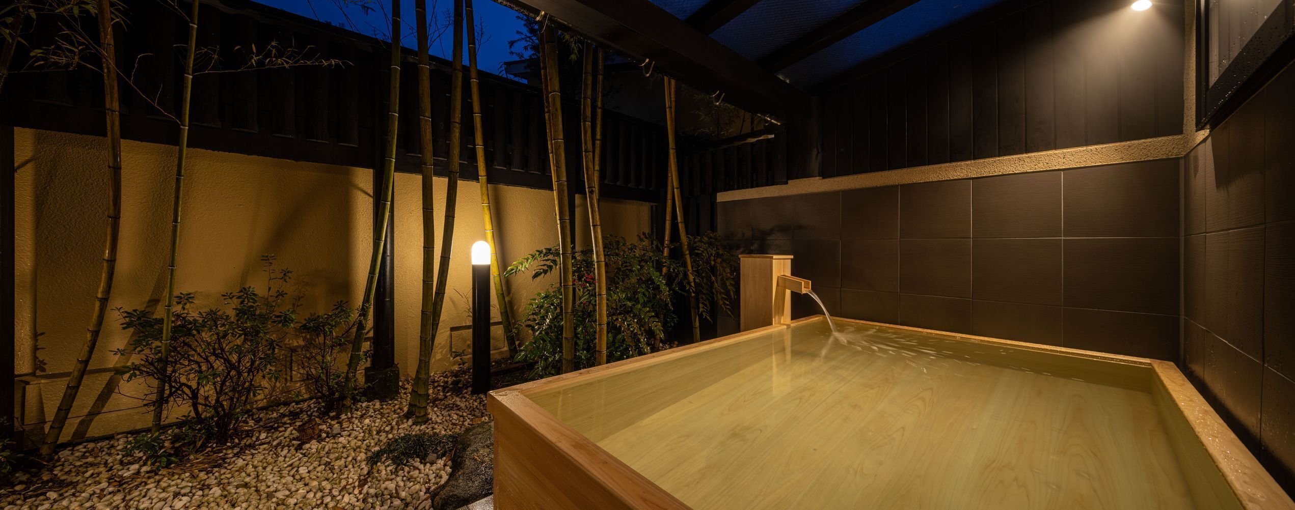 A wooden hot tub in a dimly lit outdoor area with bamboo plants and a tiled wall.