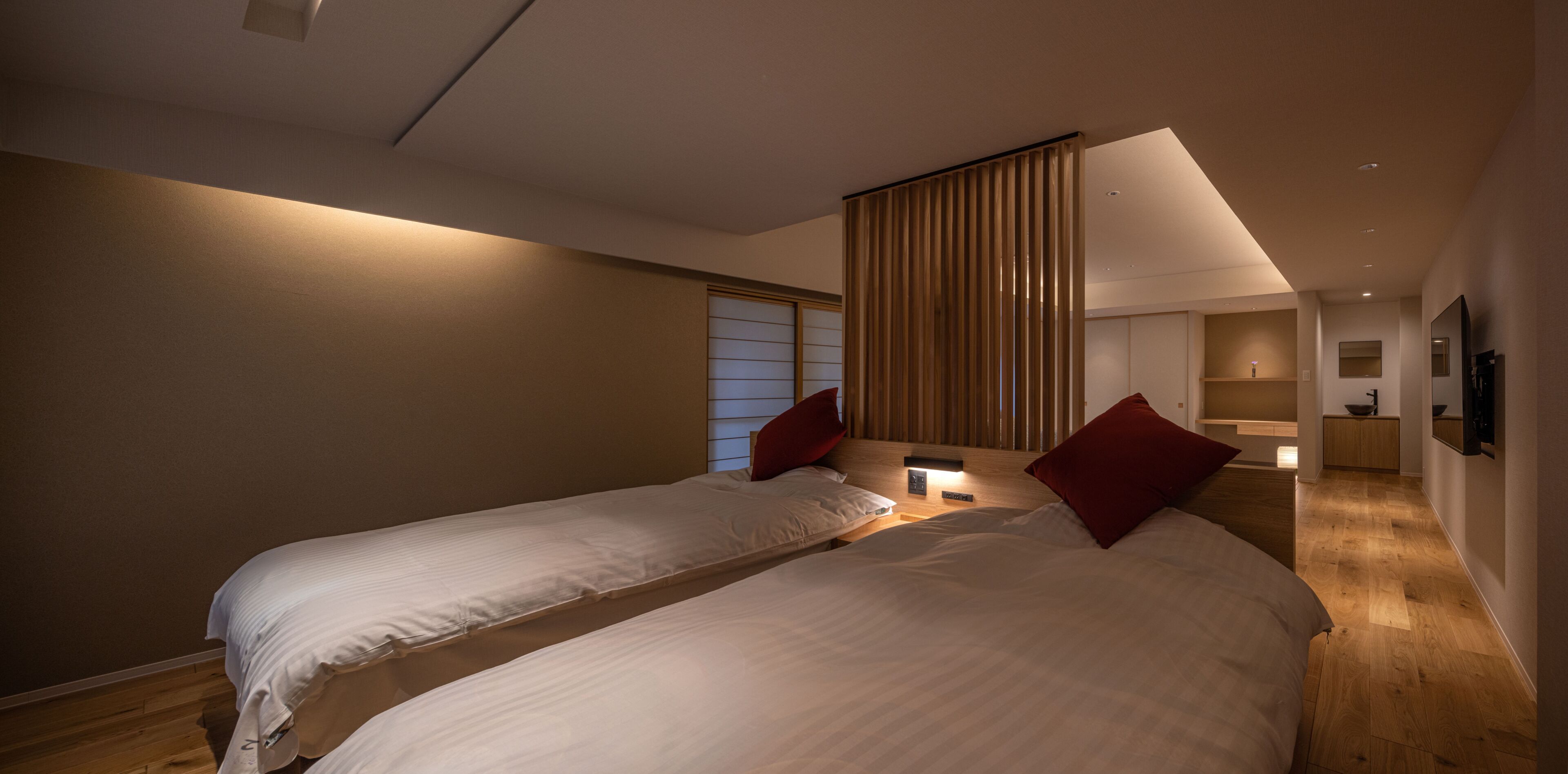 A modern hotel room with two beds, a wooden floor, a large window with blinds, a bedside table with a lamp, and a flat-screen TV mounted on the wall.
