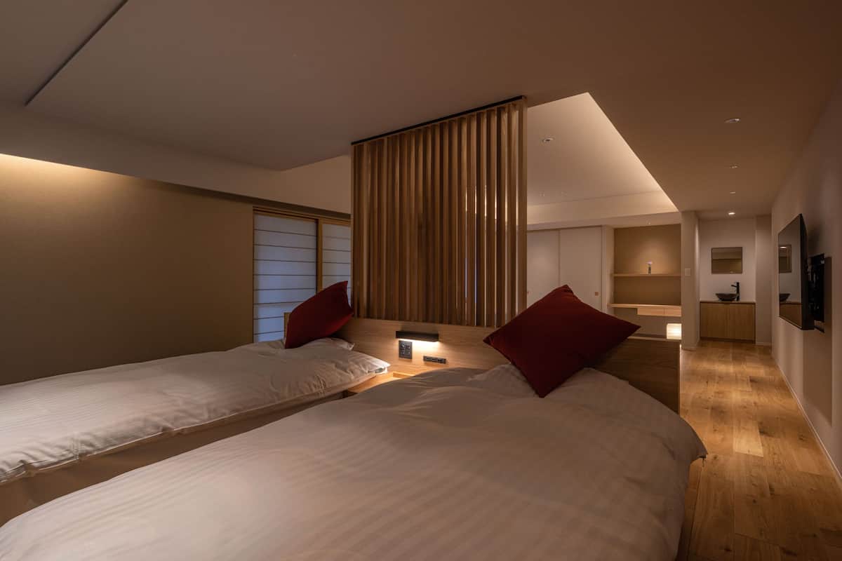 A modern hotel room with two beds, a wooden floor, a large window with blinds, a bedside table with a lamp, and a flat-screen TV mounted on the wall.