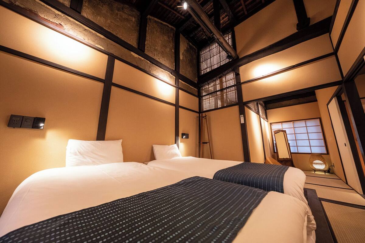 A traditional Japanese-style hotel room with tatami flooring, a large bed with white bedding, a small table, and a shoji screen.