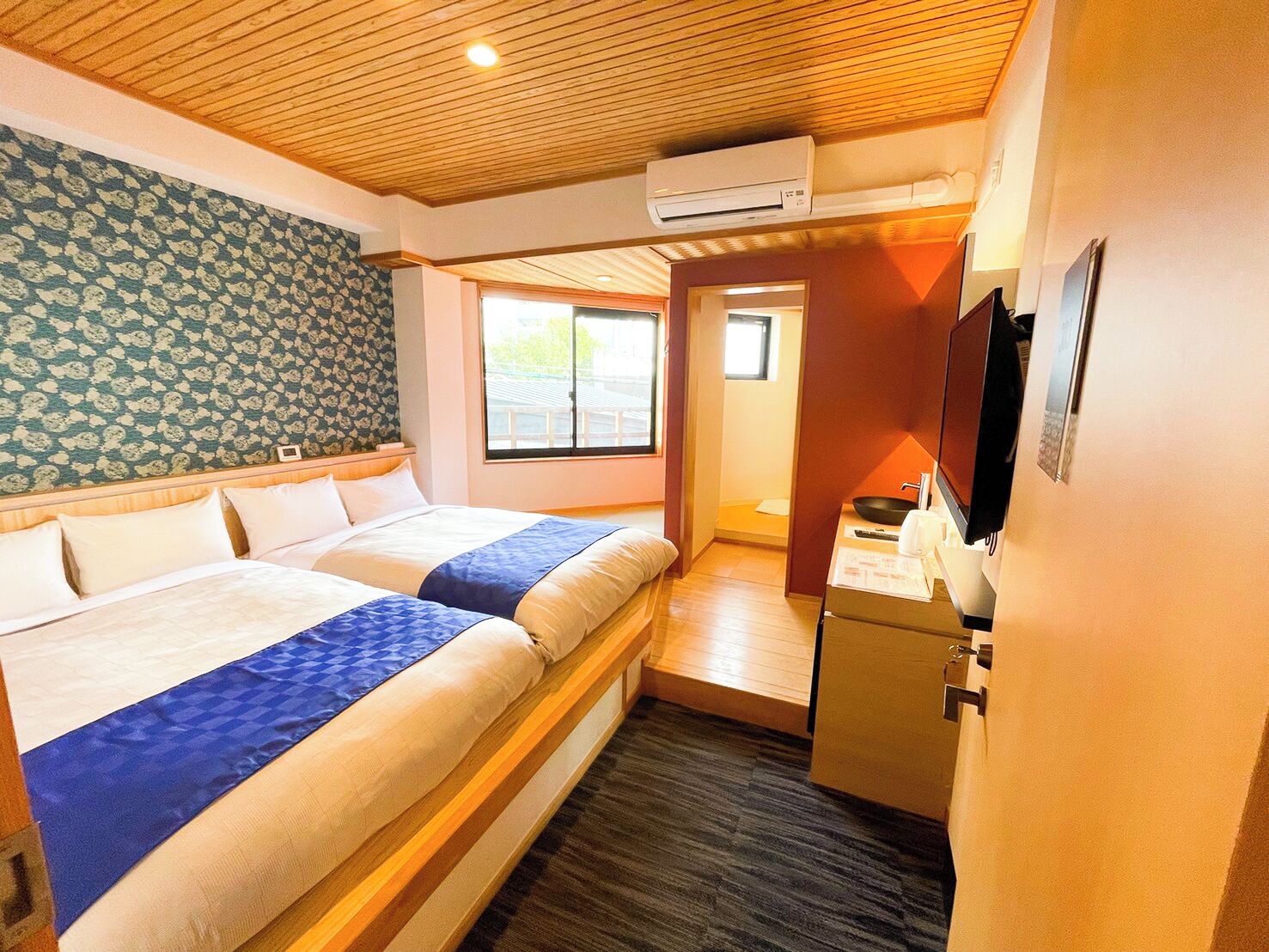 A hotel room with two beds, a wooden ceiling, an air conditioning unit, a small kitchenette, and a window.