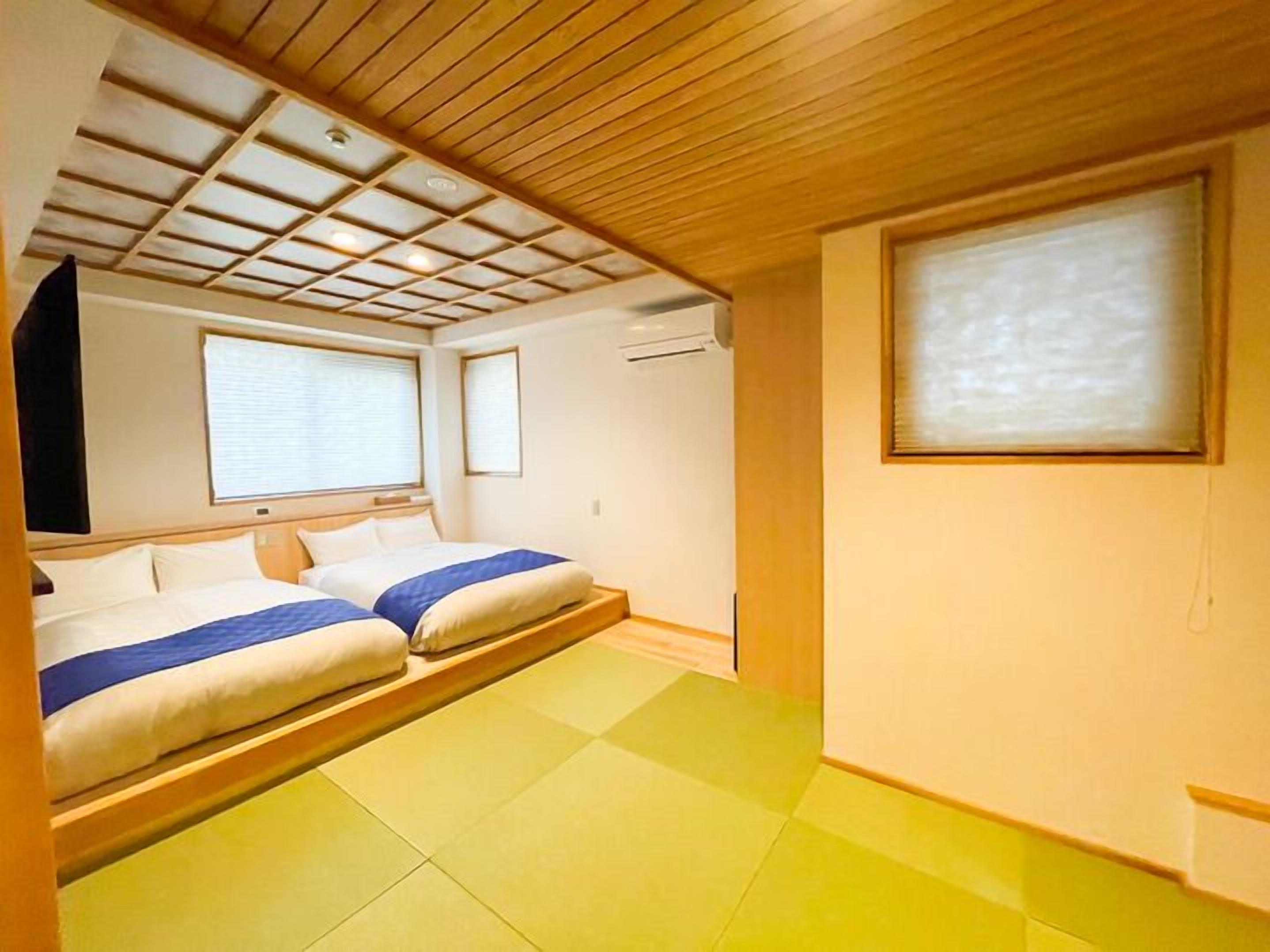A room with a wooden ceiling, two beds with white and blue bedding, a window with blinds, and a wall-mounted air conditioning unit.