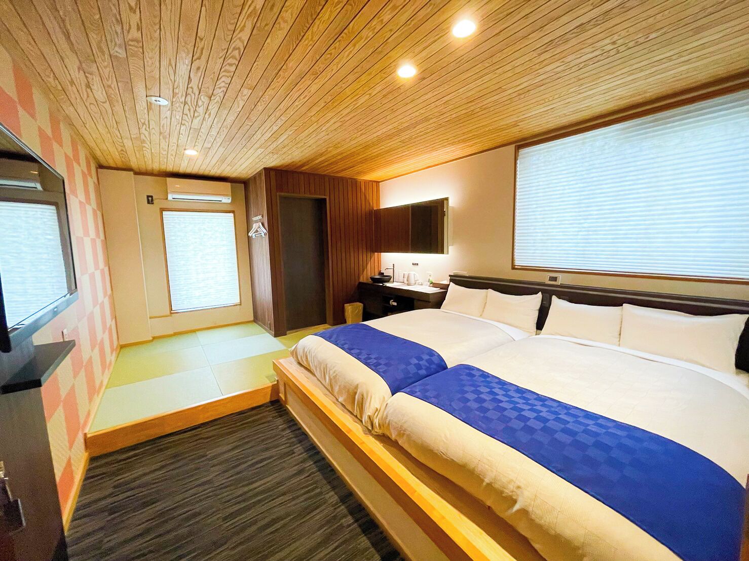 A hotel room with a wooden ceiling, a bed with blue bedding, a desk with a lamp, and a window with blinds.