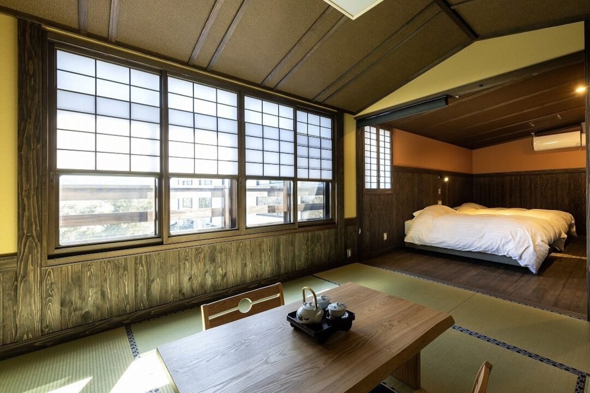 Una habitación japonesa tradicional con suelo de tatami, una cama baja, una mesa de madera con un juego de té y ventanas amplias.