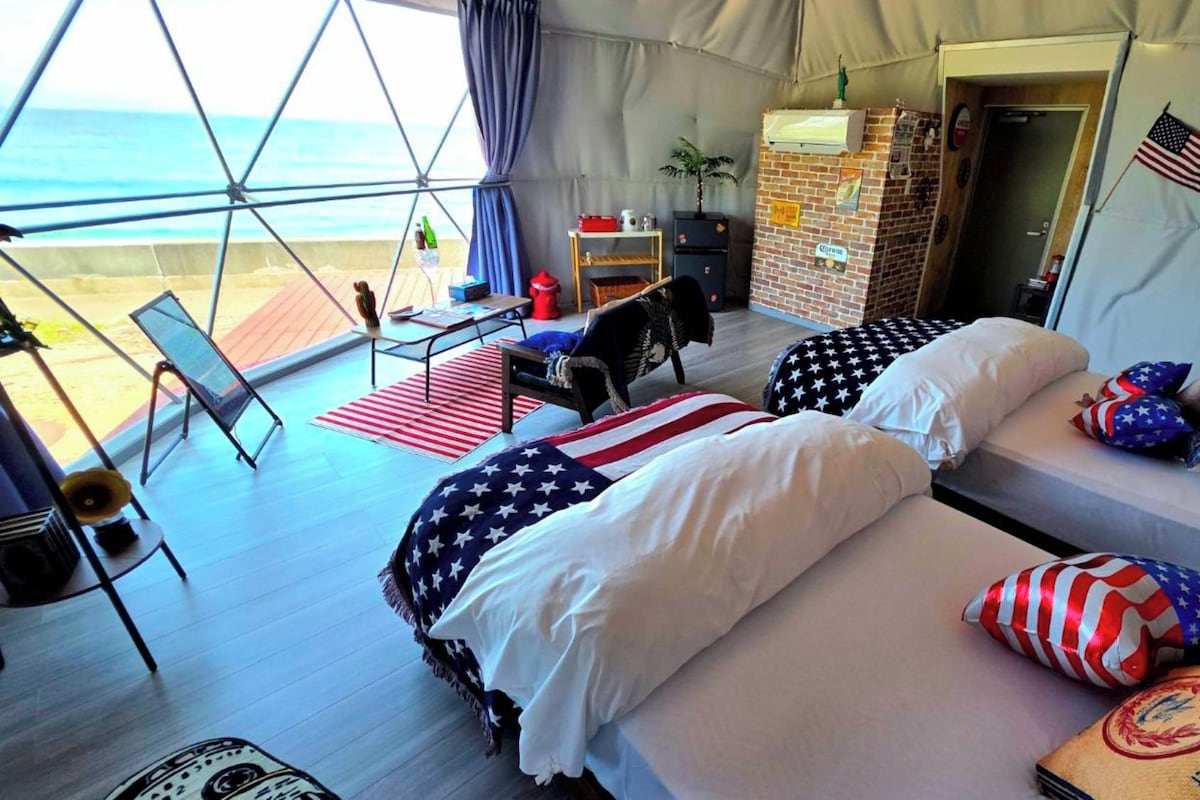 A tented accommodation with two beds, a desk, and a view of the ocean.
