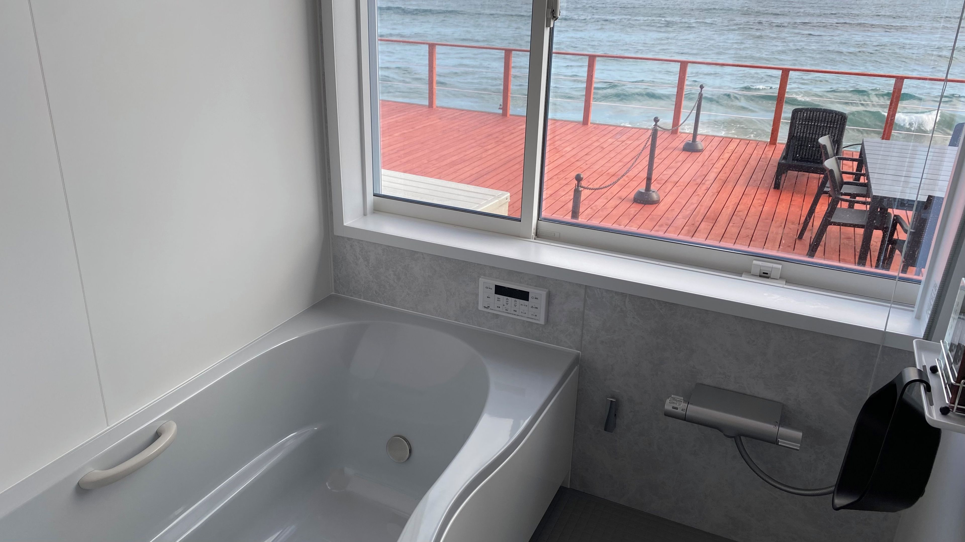 Un baño moderno con bañera, una ventana con vistas a una terraza amueblada, y un panel de control para la bañera.