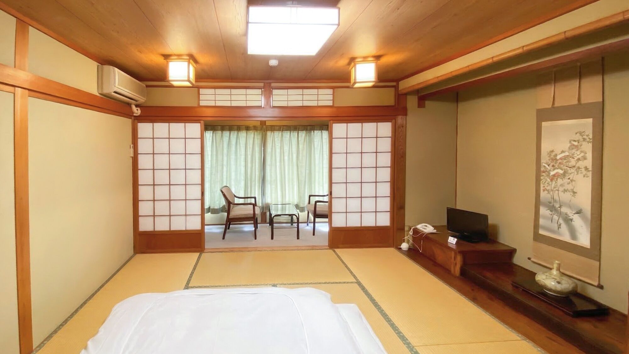Perinteinen japanilaistyylinen hotellihuone, jossa on tatamimatto, sänky, puhelimella varustettu työpöytä ja taulu seinällä.