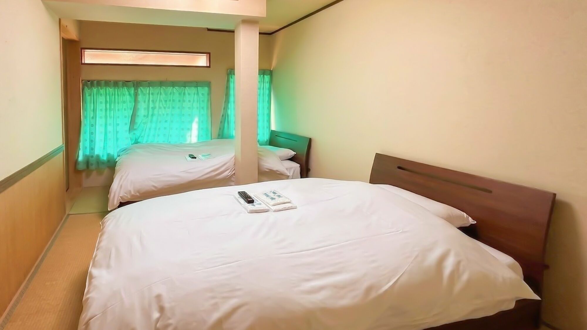 Hotellihuone, jossa on kaksi sänkyä, käytävä ja ikkuna, jossa on vihreät verhot.