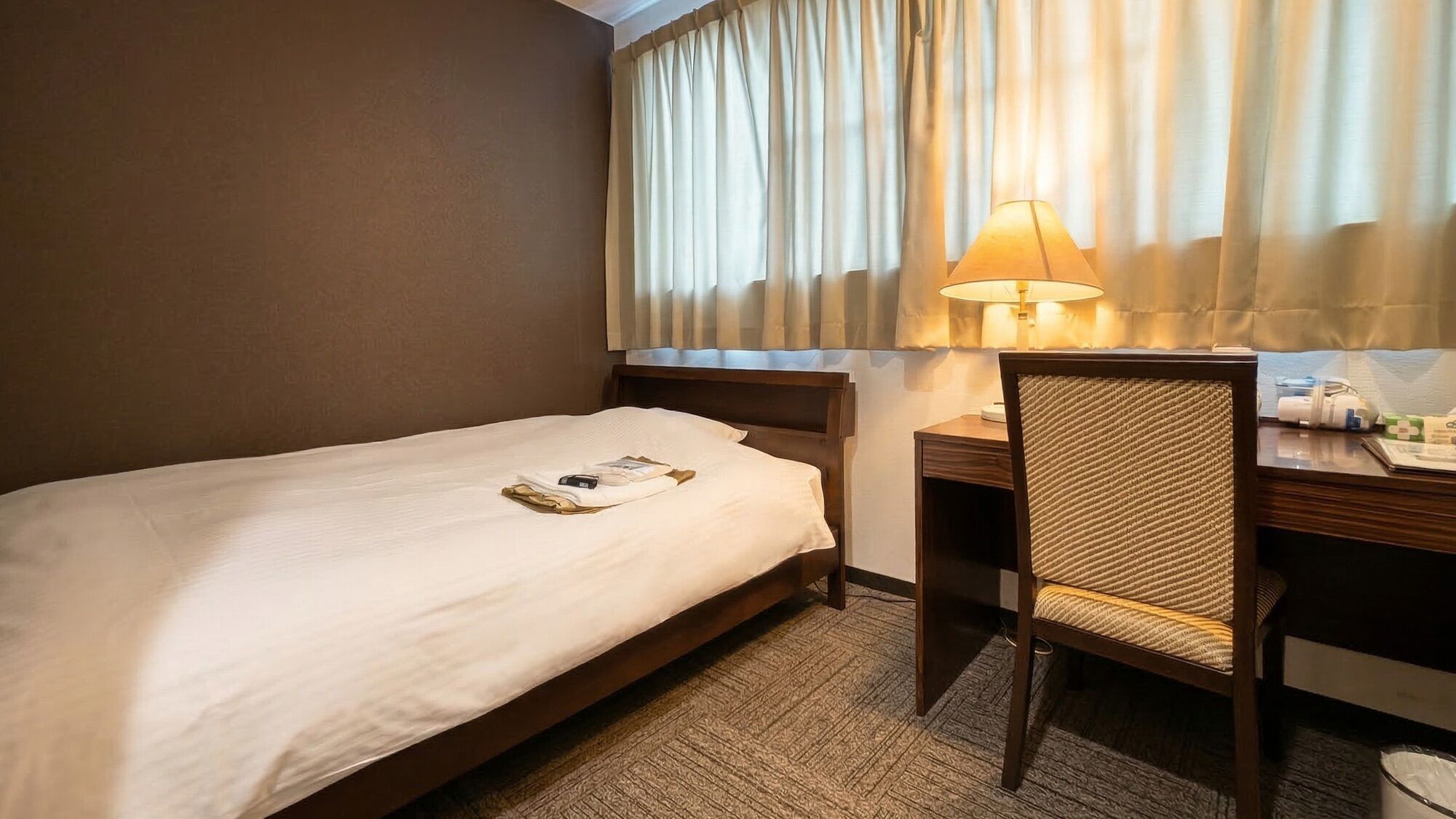 Hotellihuone, jossa on yksi sänky, työpöytä tuolilla, lamppu ja ikkuna, jossa on verhot.