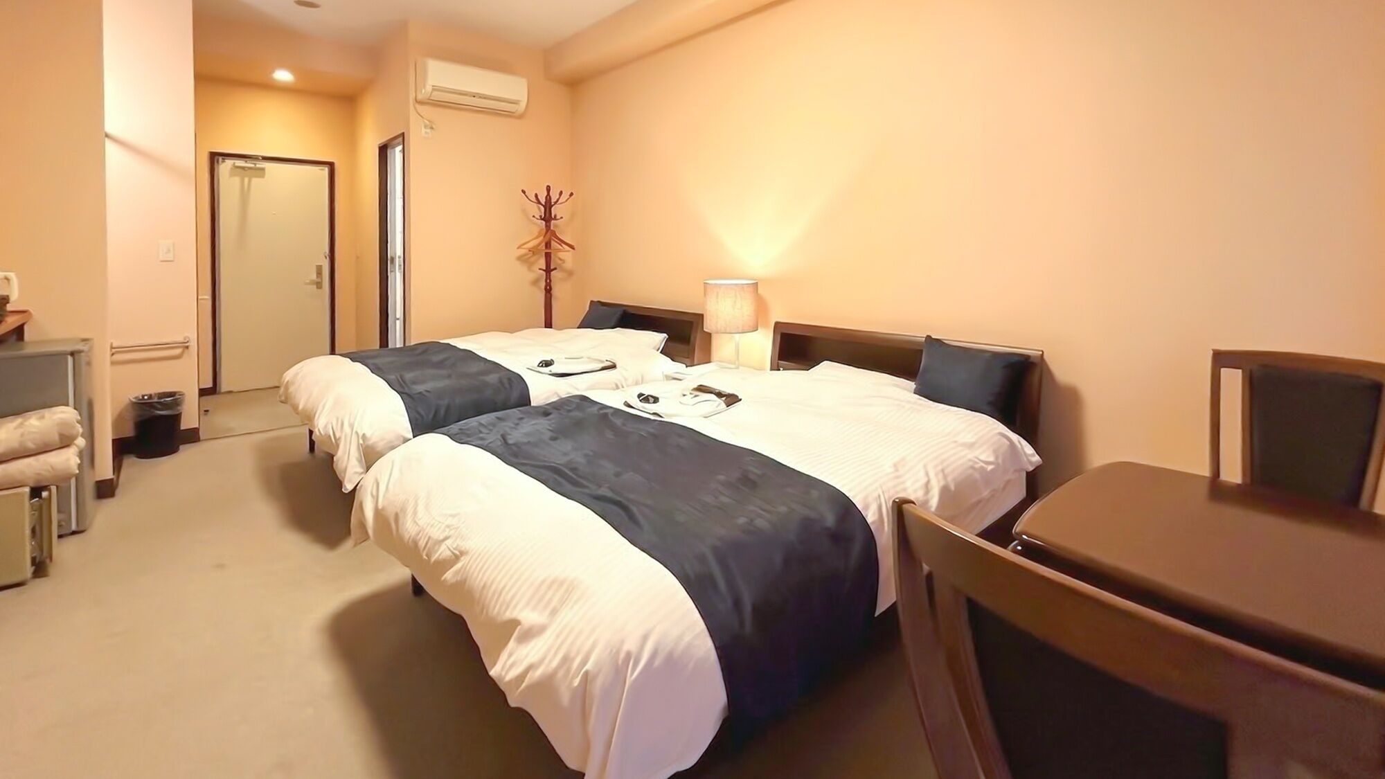 Hotellihuone, jossa on kaksi sänkyä, työpöytä, tuoli, lamppu ja naulakko.
