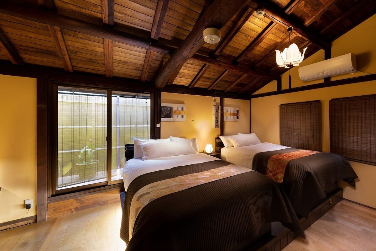 Ein Schlafzimmer mit zwei Betten, Holzbalkendecke, einem großen Fenster mit Jalousien und einer Klimaanlage.
