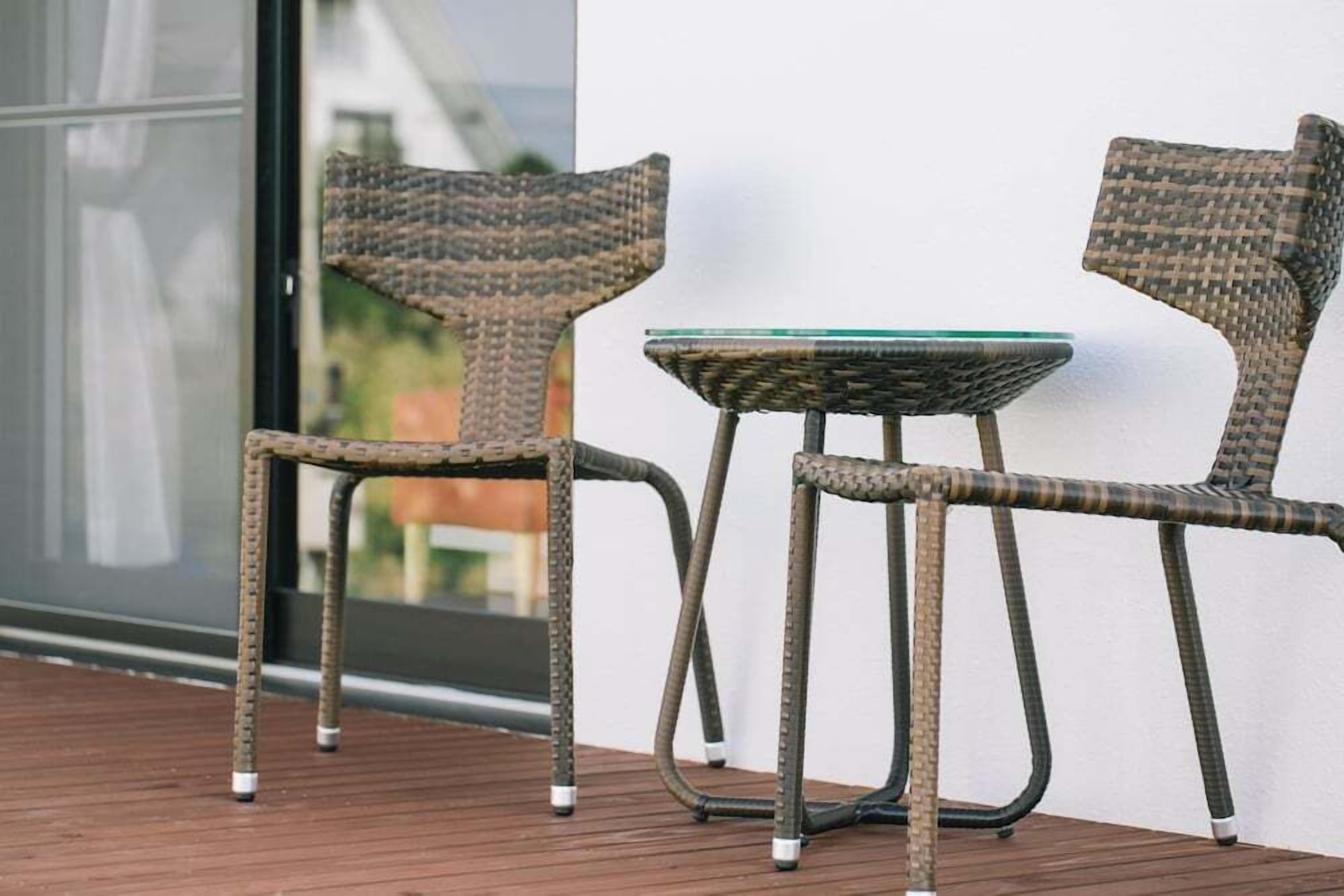 Two wicker chairs and a small glass table on a wooden deck, with a white wall and a glass door in the background.
